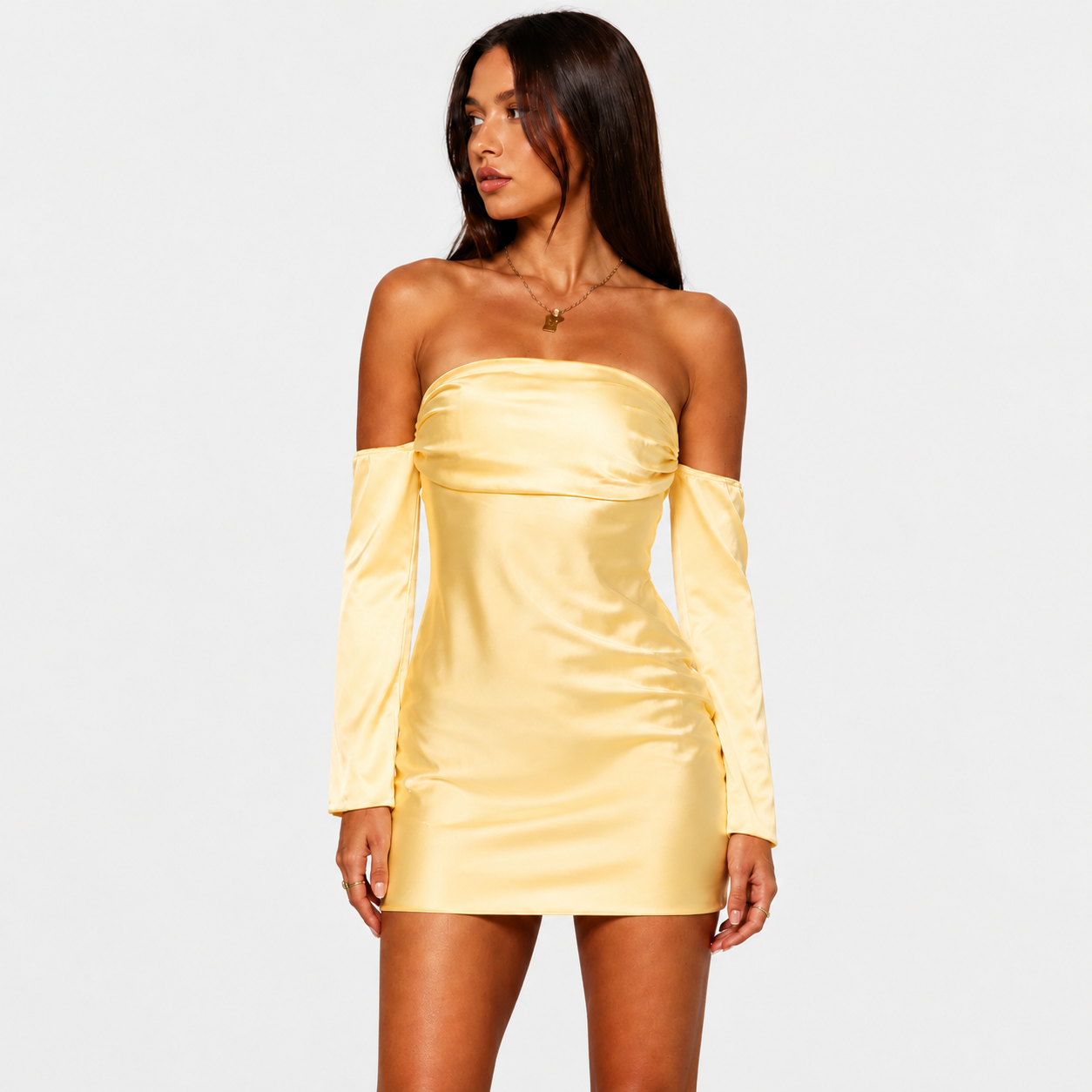 A woman posing in a yellow strapless mini dress with matching detached long sleeves, standing against a light gray background. She has long dark hair and is looking to the side while holding her hair with both hands.